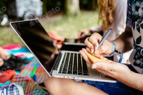 Preview: Midsection of student writing on adhesive note while sitting with laptop at park