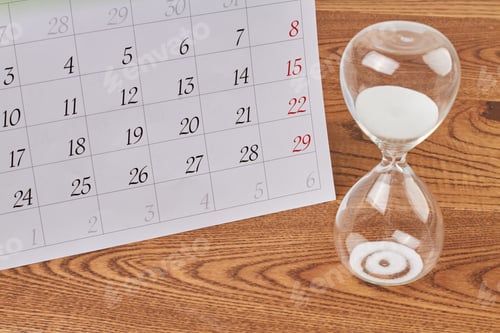 Preview: Close-up hourglass with white sand and calendar on wooden desk.
