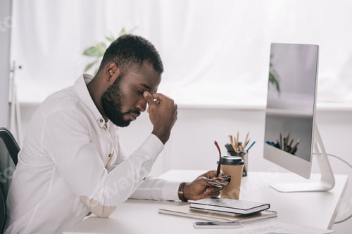Preview: side view of tired african american businessman touching nose bridge at table with computer in