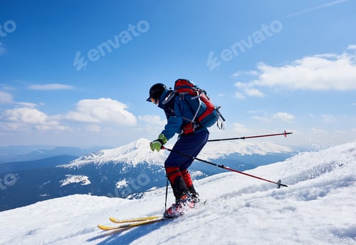 Preview: Sportsman skier in bright clothing, helmet and goggles with backpack riding down steep snowy slope.