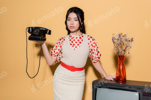 Preview: Asian model holding retro telephone near flowers on tv on orange background