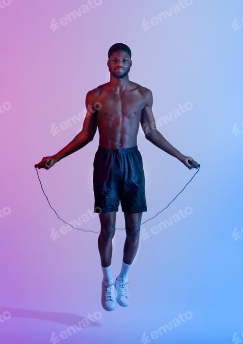 Preview: Full length portrait of handsome black sportsman with naked torso jumping on skipping rope in neon
