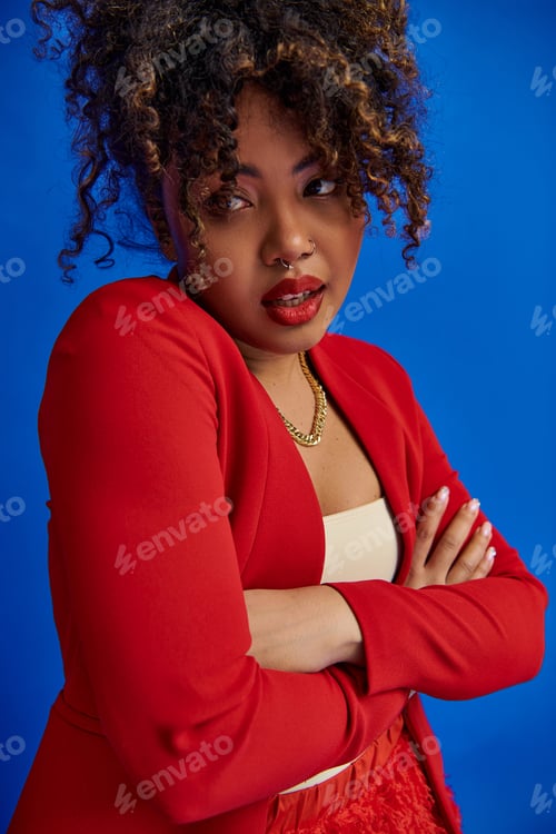 Preview: alluring trendy african american fashionista in vivid elegant outfit looking away on blue backdrop