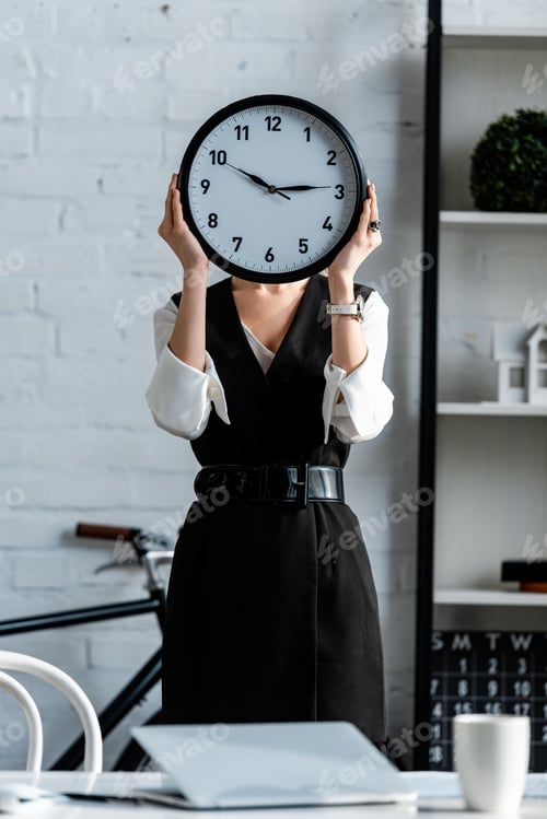 Preview: selective focus of businesswoman in formal wear holding clock in front of face at workplace