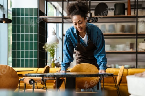 Preview: Black waitress in apron smiling while working at cafe indoors