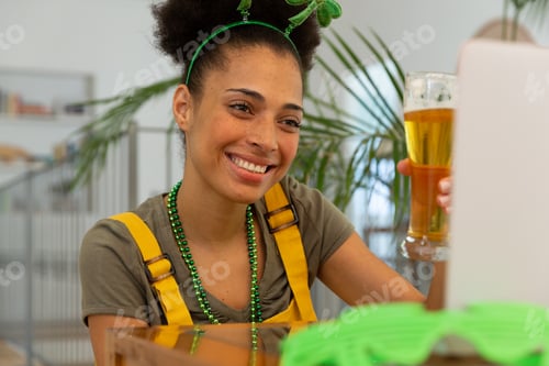 Preview: Happy mixed race woman celebrating st patrick's day making video call raising glass of beer