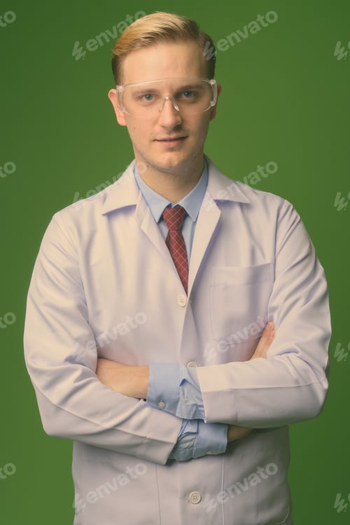 Preview: Young handsome man doctor with blond hair wearing protective glasses against green background