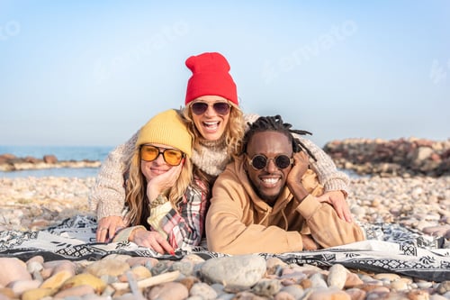 Preview: Happy multiracial diverse friends in sunglasses lying on a pebble beach smiling at camera