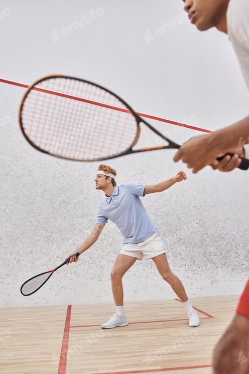 Preview: focus on redhead man in sportswear playing with african american friend inside of squash court