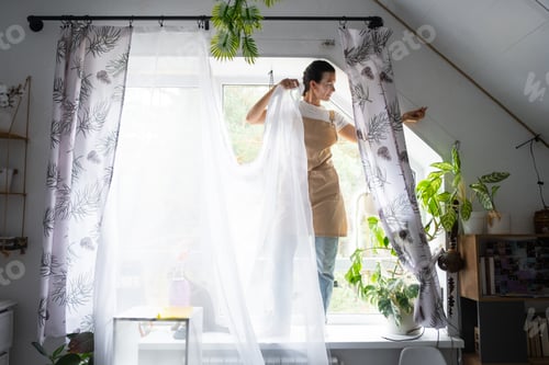 Preview: Woman in apron hangs transparent tulle curtains on large attic windows in the house inside