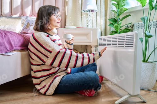 Preview: Woman warming herself in bedroom near electric heater