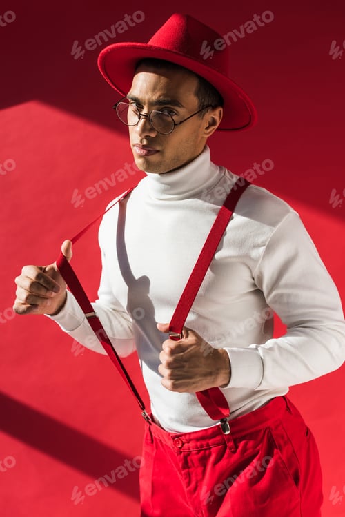Preview: fashionable mixed race man in hat and suspenders looking at camera while posing on red