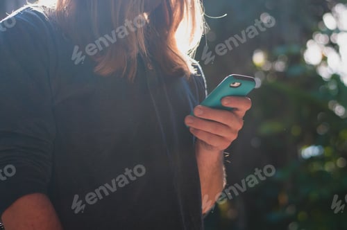 Preview: Long-haired man using his cellphone while standing in a slight beam of light.