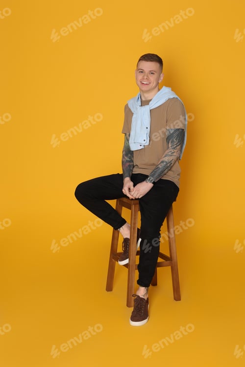 Preview: Smiling young man with tattoos sitting on stool against yellow background