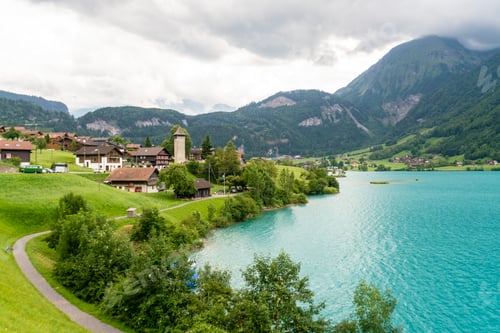 Preview: Scenic view of a quaint village by a turquoise lake with lush green hills in the background.
