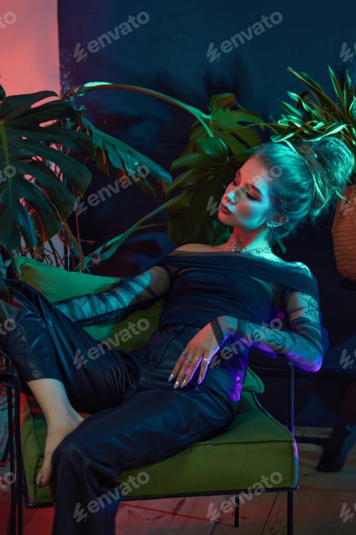 Preview: Woman sits in a green chair with her legs crossed, wearing a black off-the-shoulder top and leather