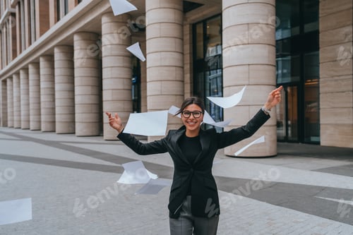 Preview: Happy brunette young entrepreneur throwing up paper sheets looks at camera happily rising hands