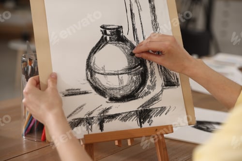 Preview: Woman drawing picture with black wax crayon on easel at wooden table in artist's studio, closeup