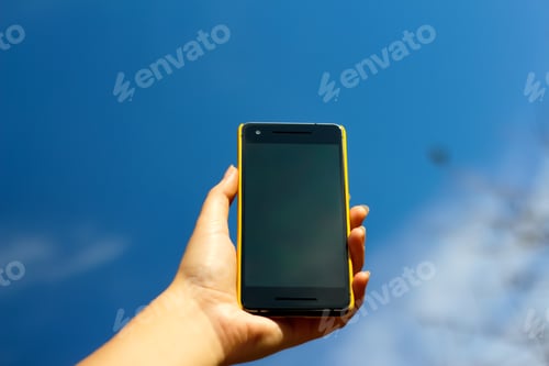 Preview: Female hand. Smart phone. Woman hand holding black smartphone in yellow plastic case on blue sky nat