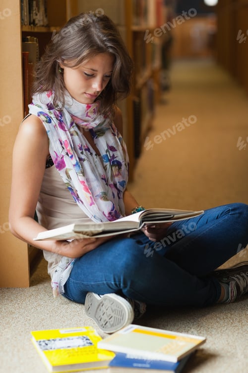 Preview: Full length of a female student sitting and reading a book in the library aisle