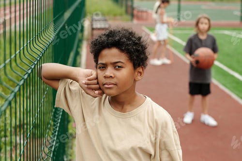 Preview: Serious cute schoolboy in t-shirt looking at camera while standing against fence