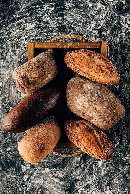 Preview: top view of arranged loafs of bread in wooden box on dark tabletop with flour
