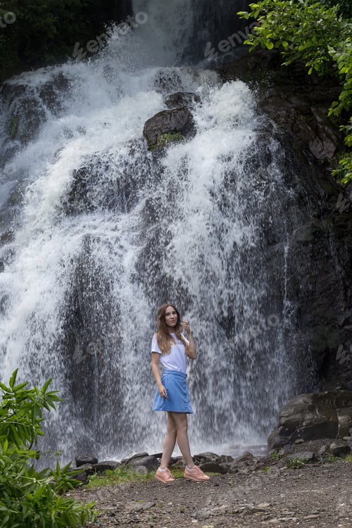 Preview: young girl in denim skirt and white t-shirt stopped by a waterfall. Gen Z