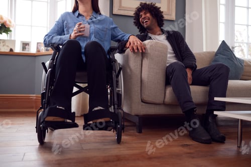 Preview: Close Up Of Couple At Home With Disabled Woman In Wheelchair Holding Hands With Man Sitting On Sofa