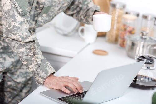 Preview: cropped view of soldier holding cup of coffee and using laptop at kitchen