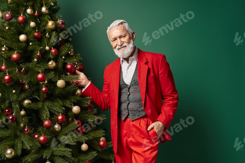 Preview: elegant stylish man dressed as Santa posing next to Christmas tree on green backdrop, winter concept