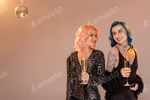 Preview: tattooed queer person with champagne glass looking at friend on beige background