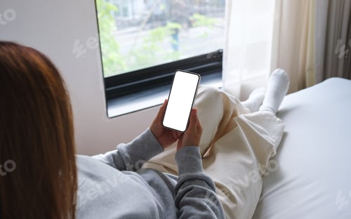 Preview: Mockup image of woman holding mobile phone with blank desktop white screen while sitting on a bed