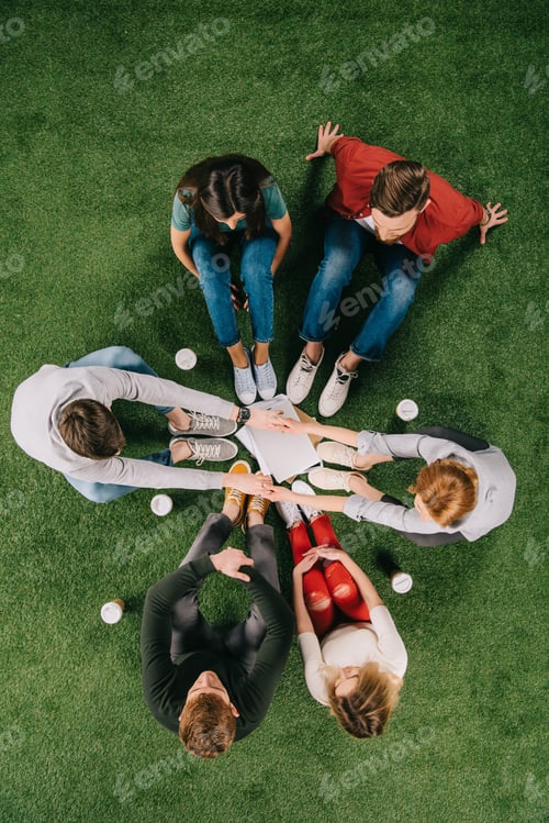 Preview: overhead view of business partners holding hands and sitting with another colleagues on grass