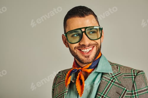 Preview: happy man in plaid jacket and bright neckerchief with sunglasses looking away on grey, vintage style