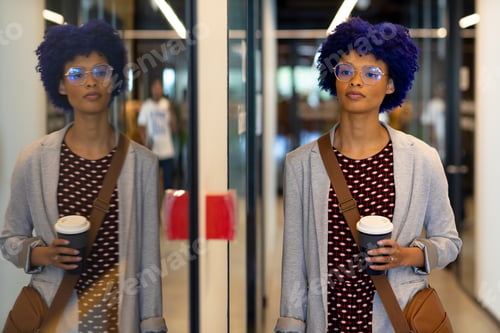 Preview: Biracial casual businesswoman with blue afro holding cup of coffee in office corridor