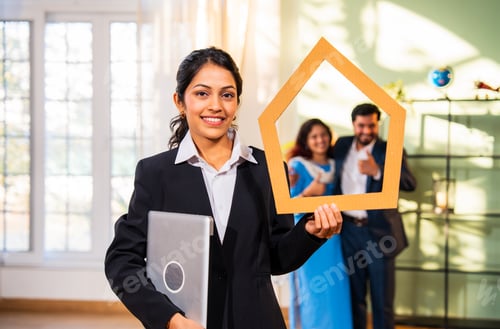 Preview: Indian woman real estate agent holding home shape cutout through which young couple buyer looks.