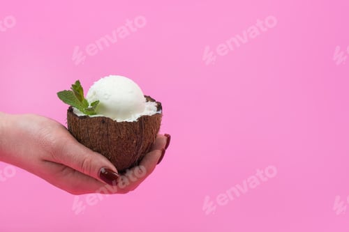 Preview: Vanilla ice cream balls in a fresh coconut half, decorated with mint leaves in your hand on a pink