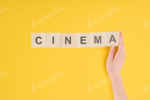 Preview: top view of female hand touching wooden cubes with 'cinema' lettering isolated on yellow
