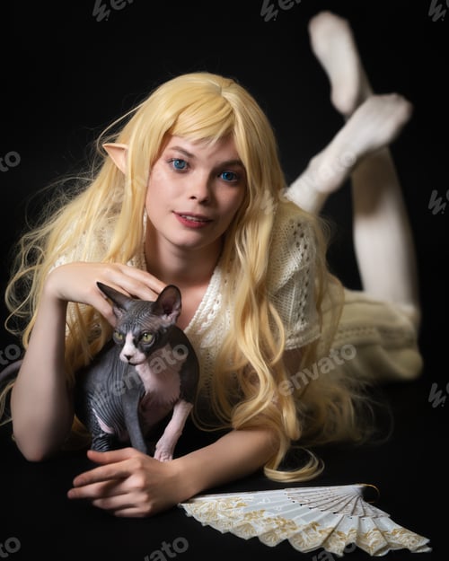 Preview: Playful blue eyed blonde female elf cosplayer lying down on stomach and stroking Sphinx kitten