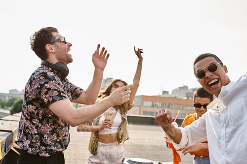 Visualização: amigos multiculturais alegres com óculos de sol estilosos festejando juntos com DJ no terraço