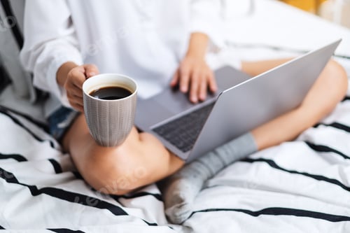 Vorschau: eine Frau benutzt und arbeitet am Laptop, trinkt Kaffee, während sie auf einem weißen, gemütlichen Bett sitzt