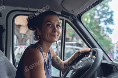 Preview: Young woman, with tattoos and piercings, driving a van, looks at the room with satisfied expression.