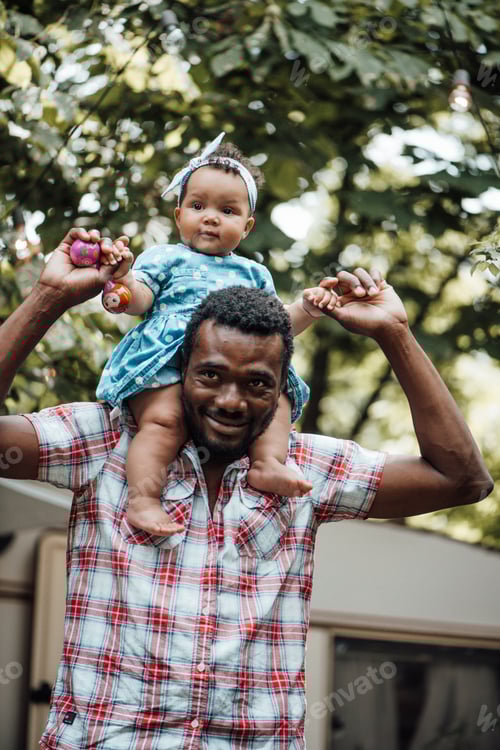 Preview: Happy black ethnicity dad carries toddler baby girl on his neck outdoor next to retro caravan house