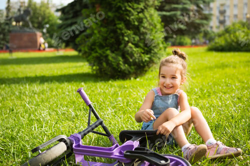 Preview: Cheerful girl sitting resting on the grass in a park near to the purple running bike