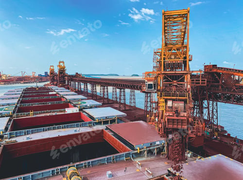 Preview: Iron ore conveyor loading cargo in the bulk carrier with a cloudy blue sky in the background