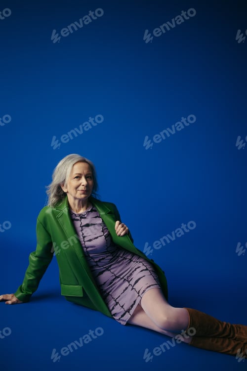 Preview: fashionable senior woman in green leather jacket over purple dress smiling at camera while sitting