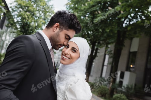 Preview: groom in suit hugging cheerful muslim bride in hijab and white dress