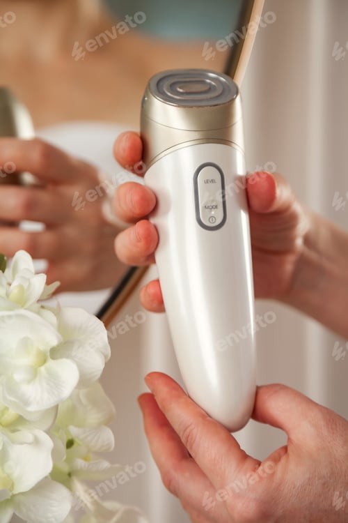 Preview: Close-up hands of middle age lady holding anti-aging electric massager doing therapy facial massage