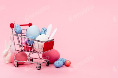 Vorschau: Bunte Ostereier und dekorative Hasen im Einkaufswagen auf rosafarbenem Hintergrund