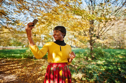Preview: African american girl at yellow and red dress at golden autumn fall park making selfie on phone.